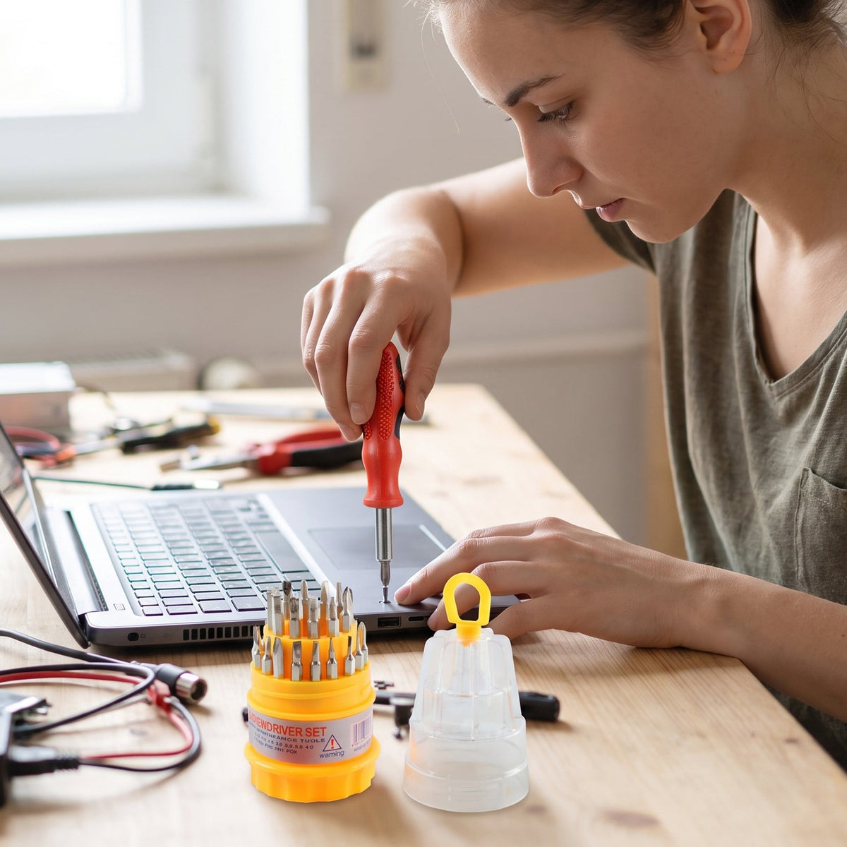 A person uses a red and black screwdriver to work on a laptop, with the yellow and clear screwdriver set on a wooden table.