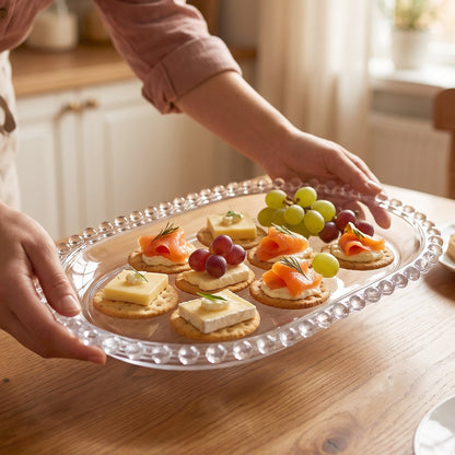 A person's hands present a clear oval glass tray filled with crackers topped with cheese, salmon, and grapes.