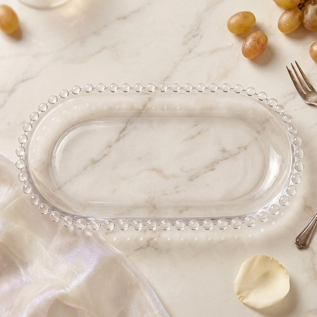 An empty clear oval glass tray with a beaded border sits on a marble surface with a white fabric and fork.
