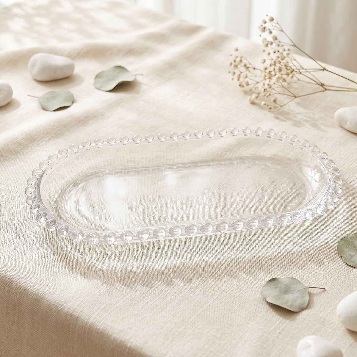 An empty clear oval glass serving tray with a beaded edge rests on a light linen tablecloth with decorative leaves.
