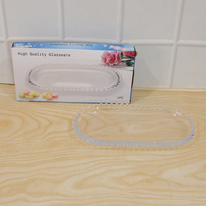 A clear oval glass serving tray is displayed next to its retail packaging on a wooden surface.