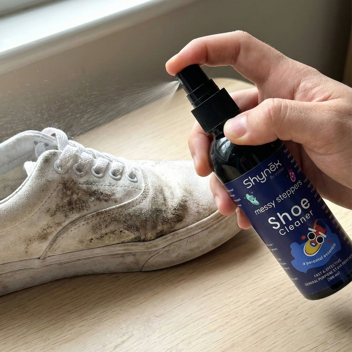 A hand sprays Shynex shoe cleaner onto a heavily soiled white canvas shoe near a window.
