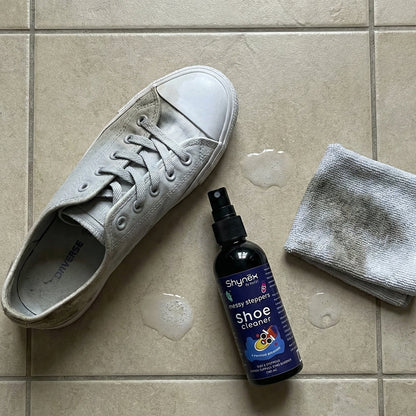 A dirty grey Converse shoe, a bottle of Shynex cleaner, and a soiled grey cloth lie on a tiled floor.
