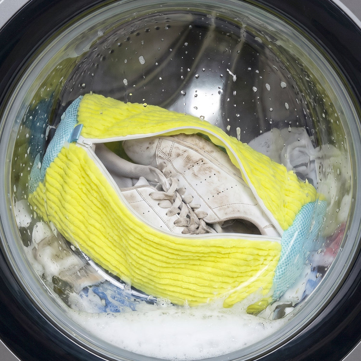 A yellow and blue shoe laundry bag with dirty white shoes inside, tumbling in a washing machine with soapy water.