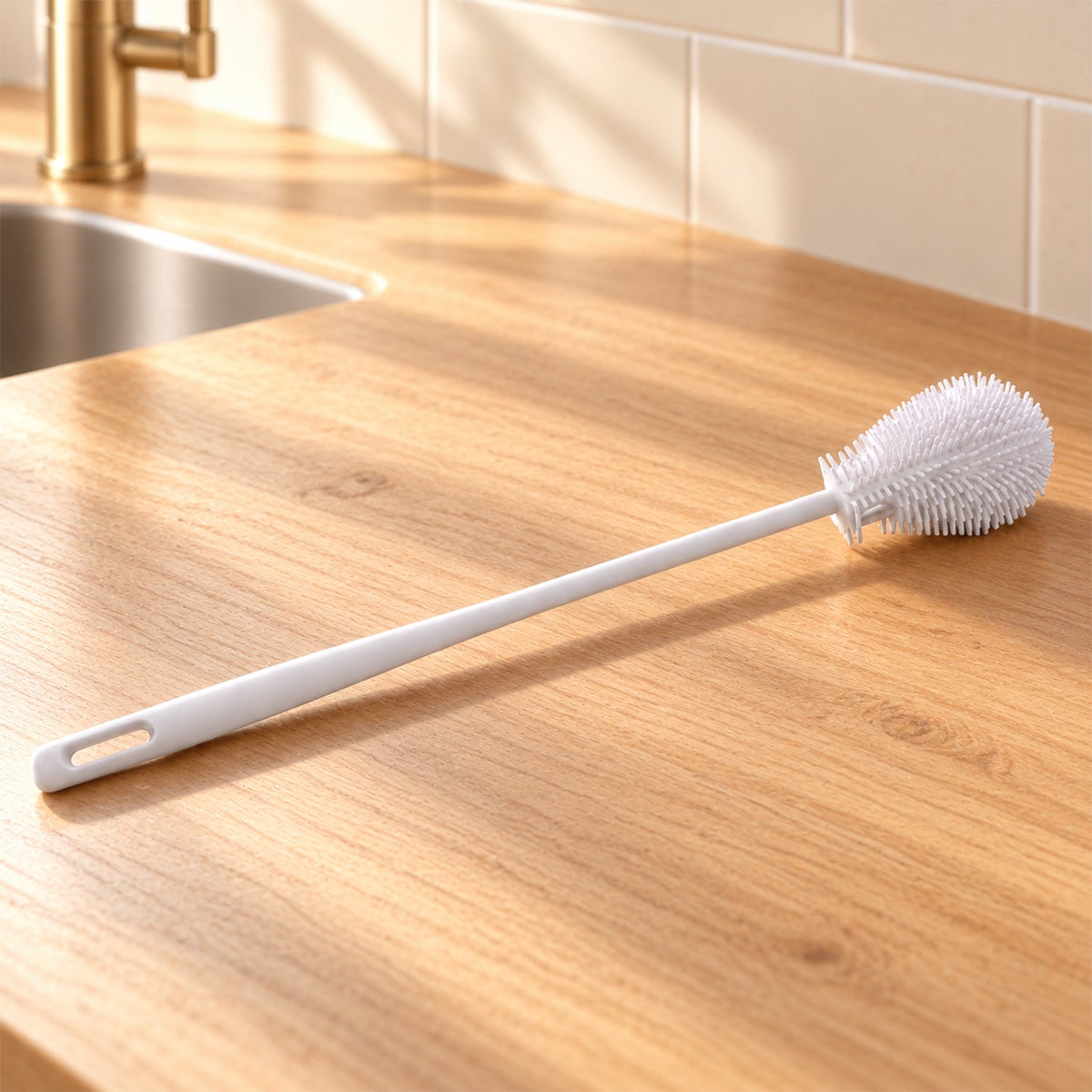 A white silicone bottle cleaning brush rests on a light wooden countertop next to a kitchen sink with a gold faucet.