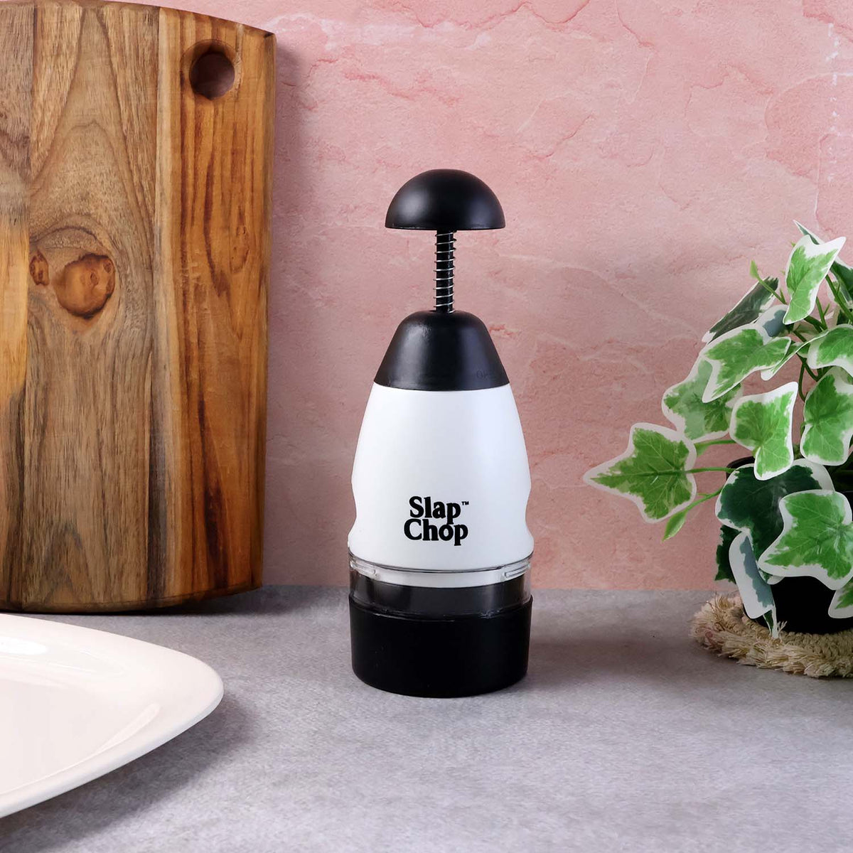 The white and black manual food chopper stands upright on a gray surface, beside a wooden board and a green potted plant.