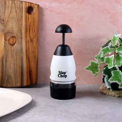 The white and black manual food chopper stands upright on a gray surface, beside a wooden board and a green potted plant.