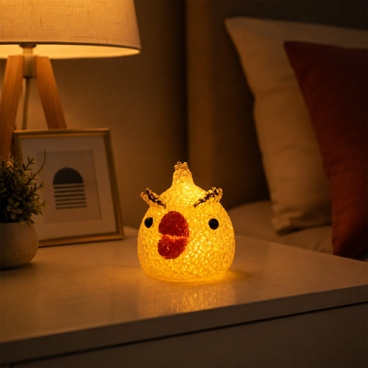 A glowing yellow chick-shaped night lamp with a red beak and black eyes sits on a white nightstand next to a lamp.