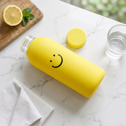 A yellow glass water bottle with a smiley face lies on a marble counter next to a lemon, mint, and glass of water.