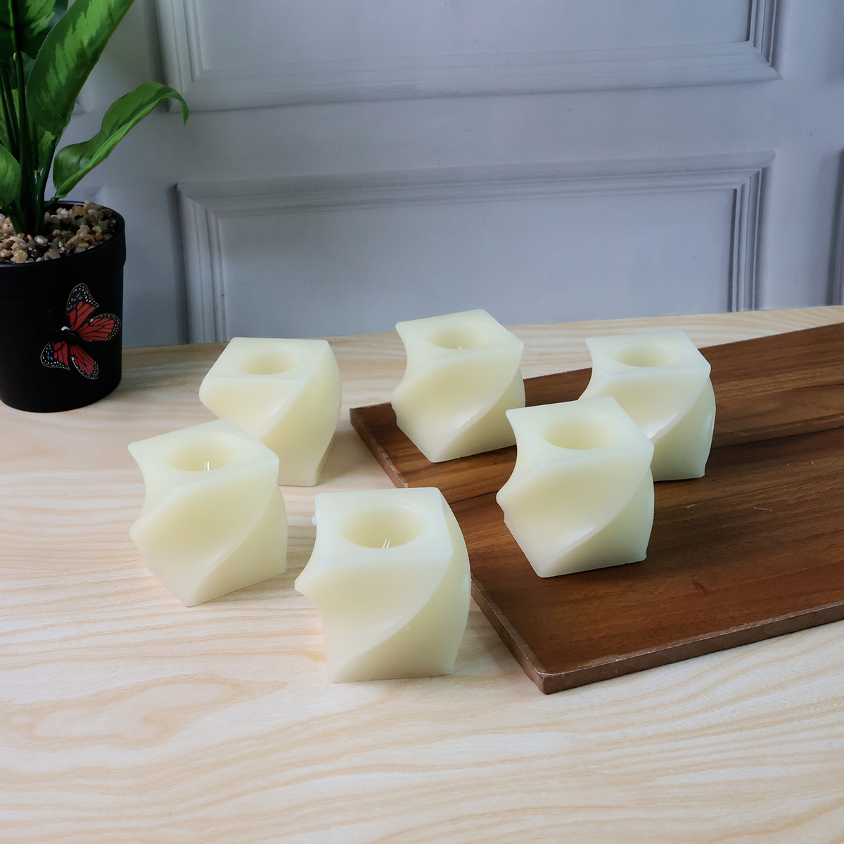 Six unlit cream-colored flameless LED candles with a twisted design are arranged on a light wooden surface next to a plant.