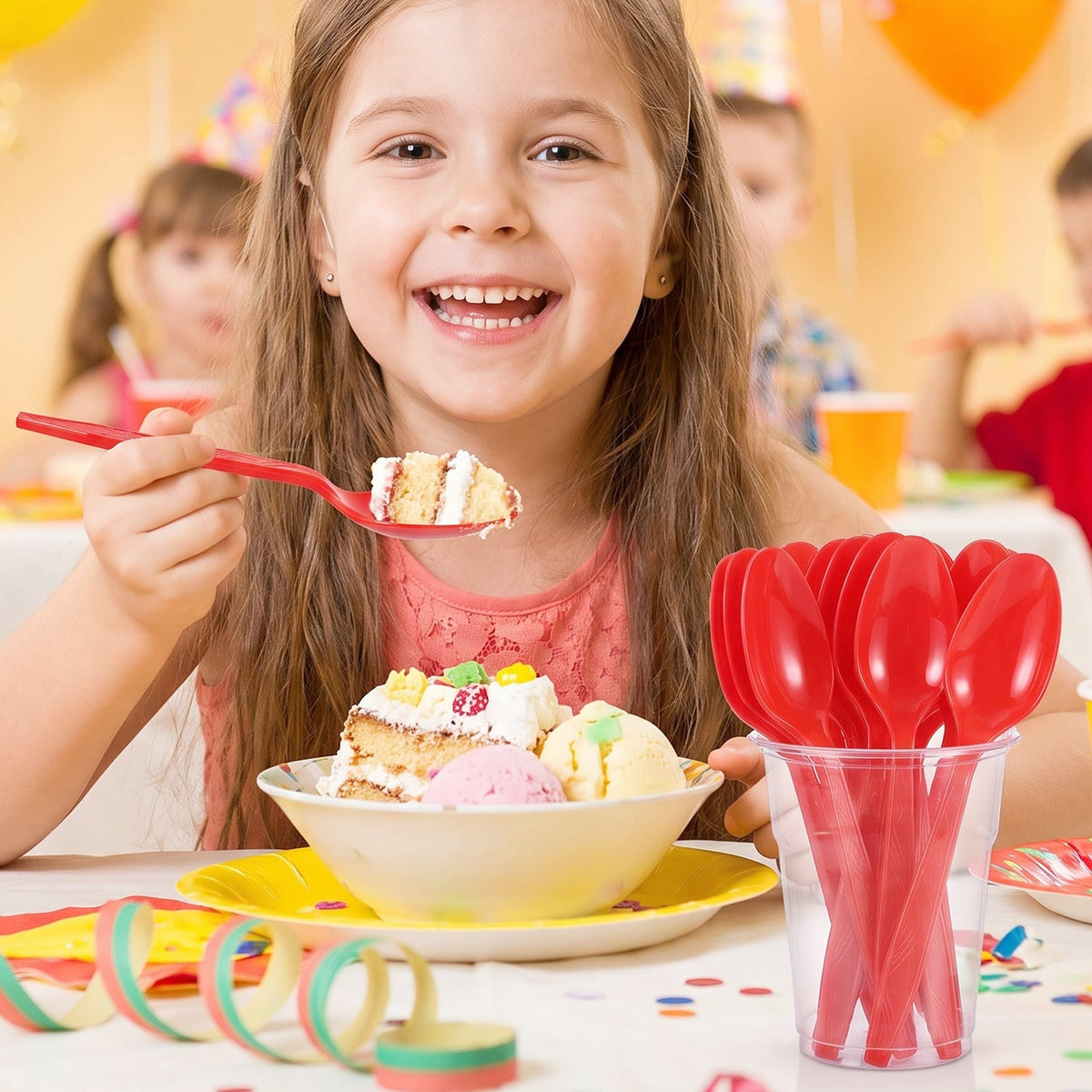 A smiling girl at a party holds a red plastic spoon with cake, with a cup of spoons nearby.