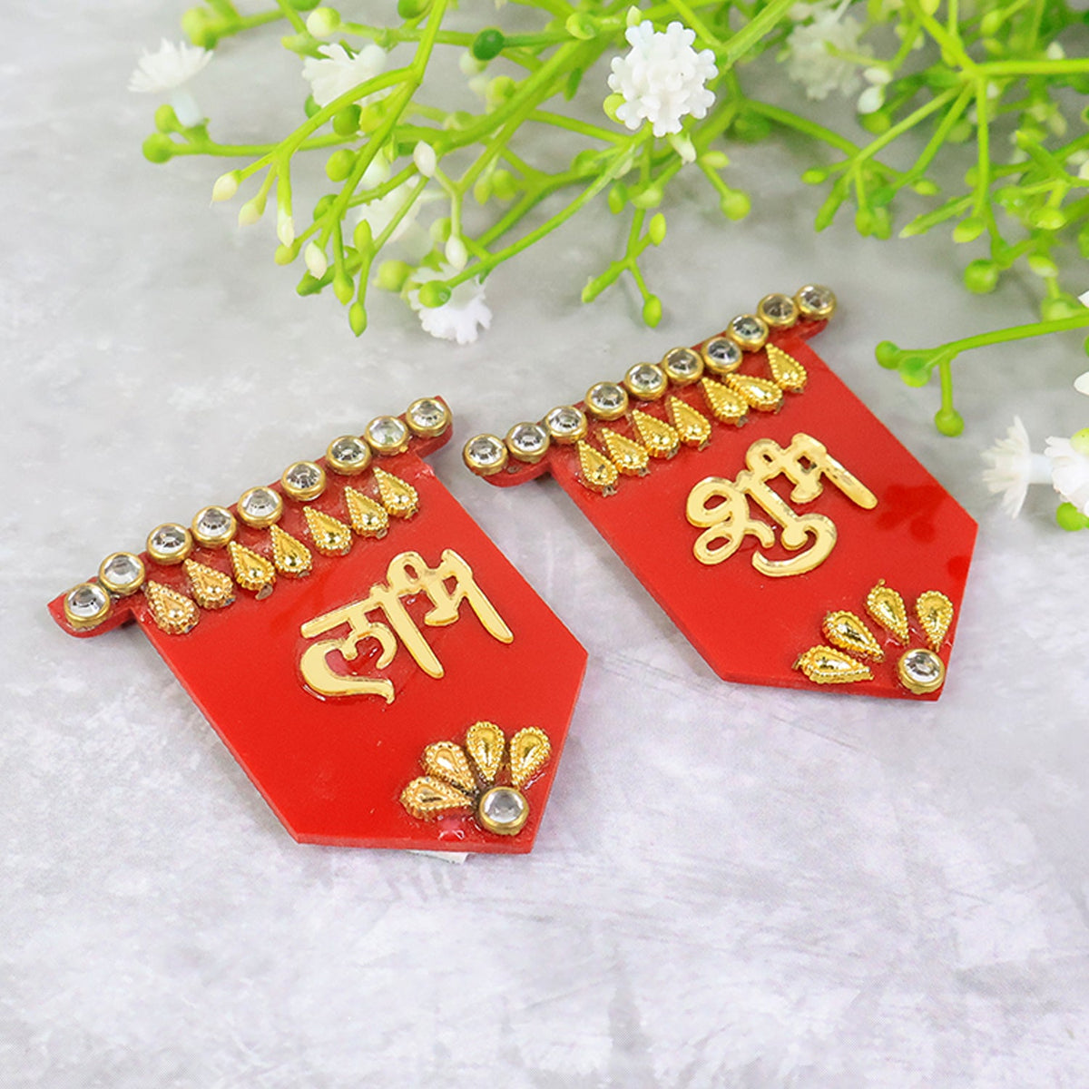 Two red Shubh Labh wall hangings with gold and clear stone embellishments, angled on a grey surface with green and white