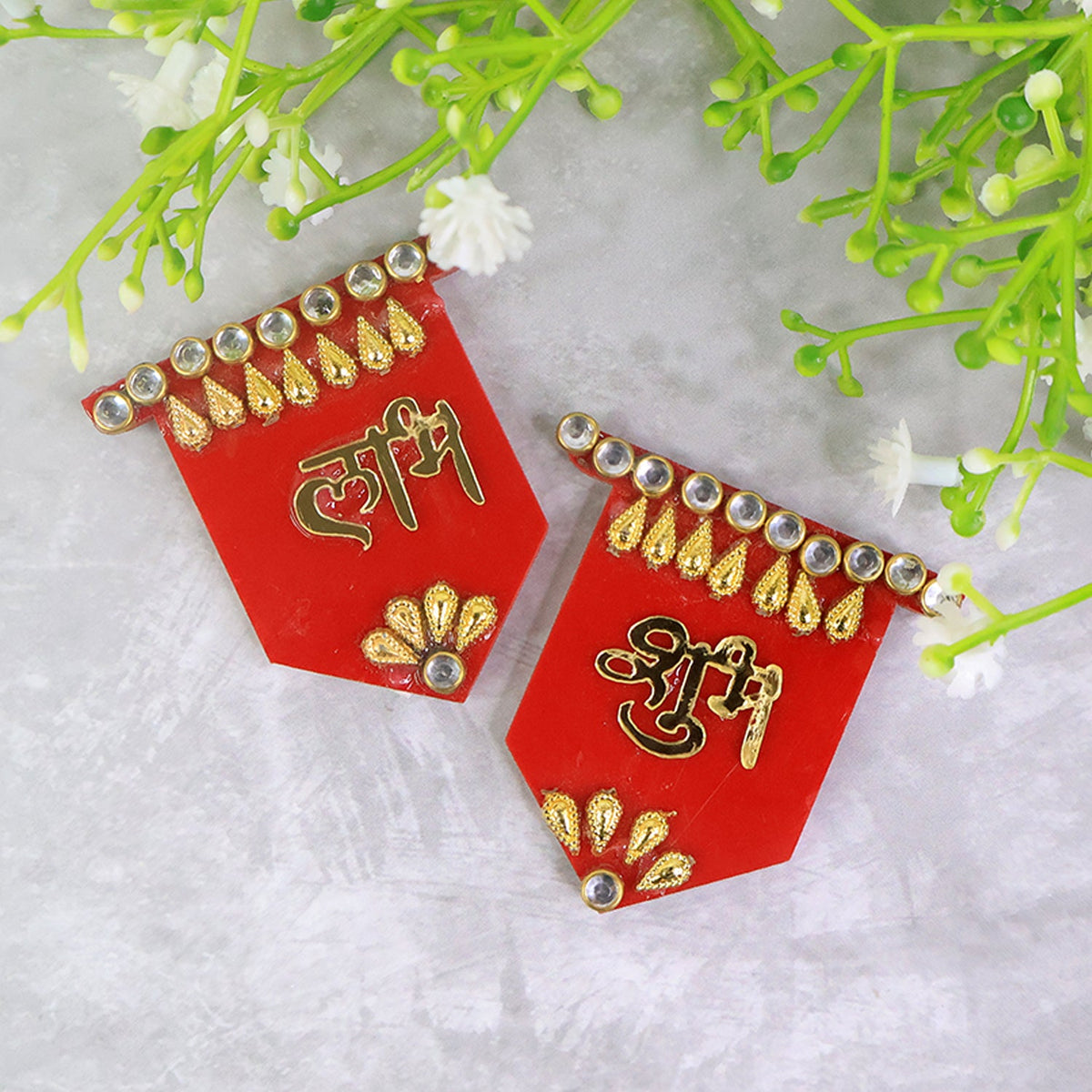 Two red Shubh Labh wall hangings with gold and clear stone embellishments, positioned on a grey textured surface with green