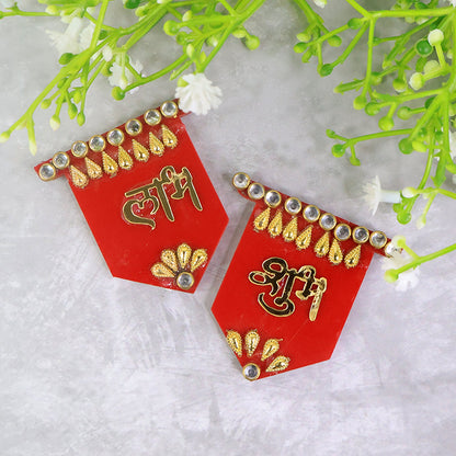 Two red Shubh Labh wall hangings with gold and clear stone embellishments, positioned on a grey textured surface with green