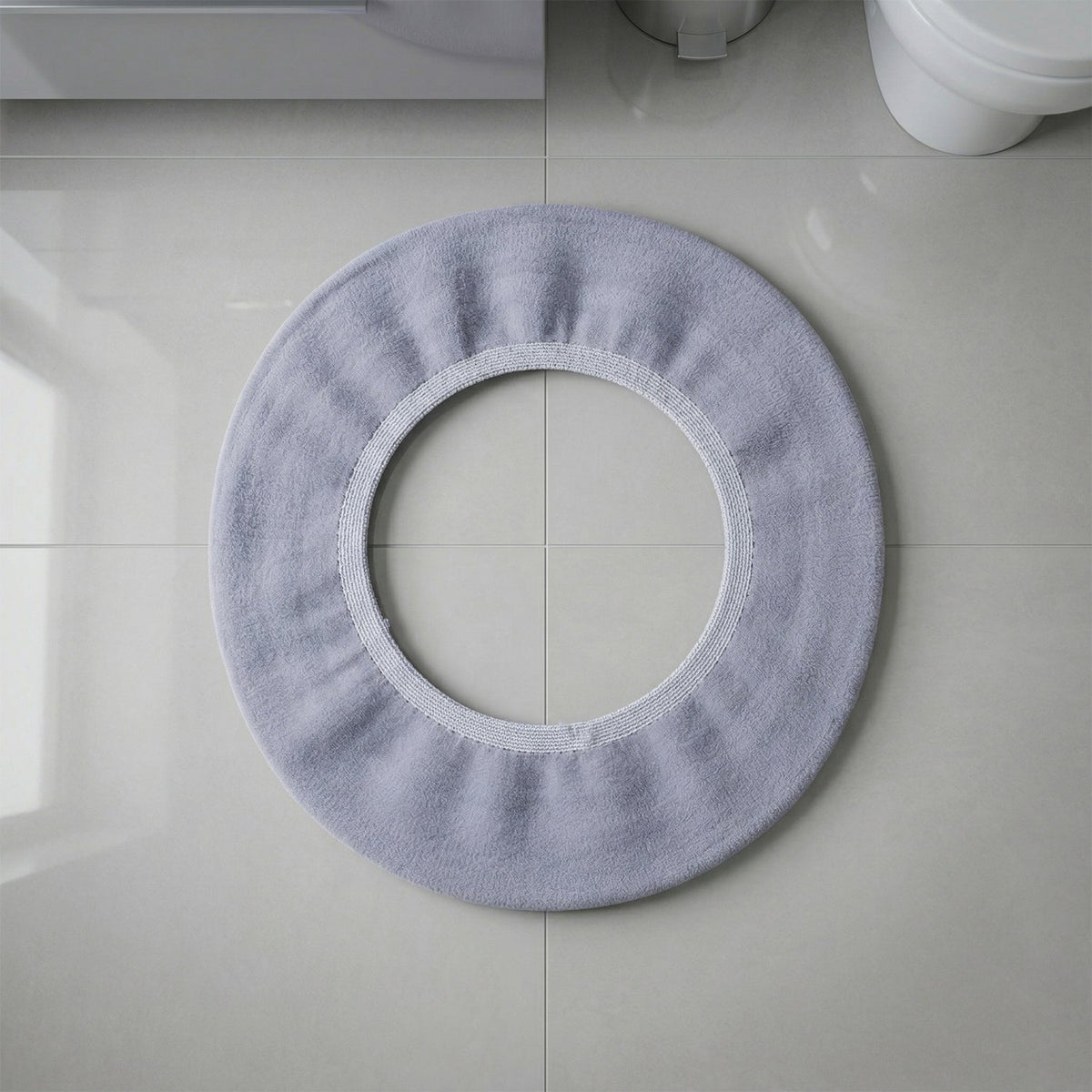 A light grey elastic toilet seat cover laid flat on light grey tiled flooring.
