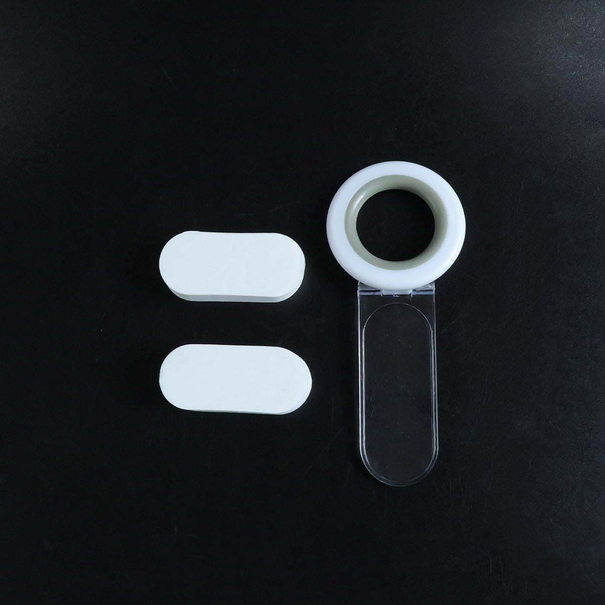 A white and gray toilet seat lifter with a clear handle and two white oval adhesive pads arranged on a dark background.