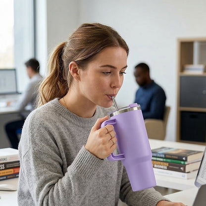 A woman sips from a purple insulated tumbler with a straw while working on a laptop at a desk.