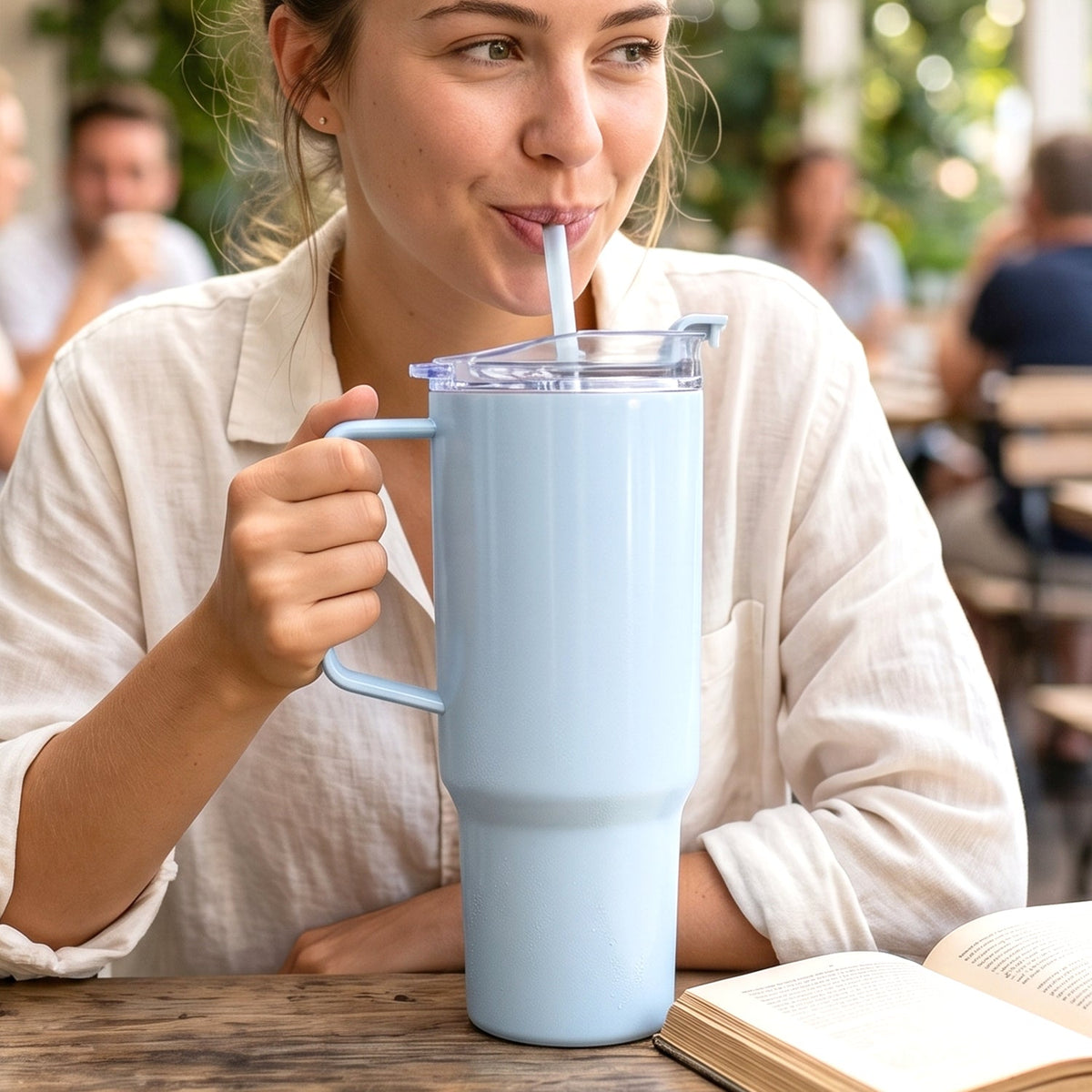 A woman sips from a large light blue handled tumbler with a straw at an outdoor cafe.