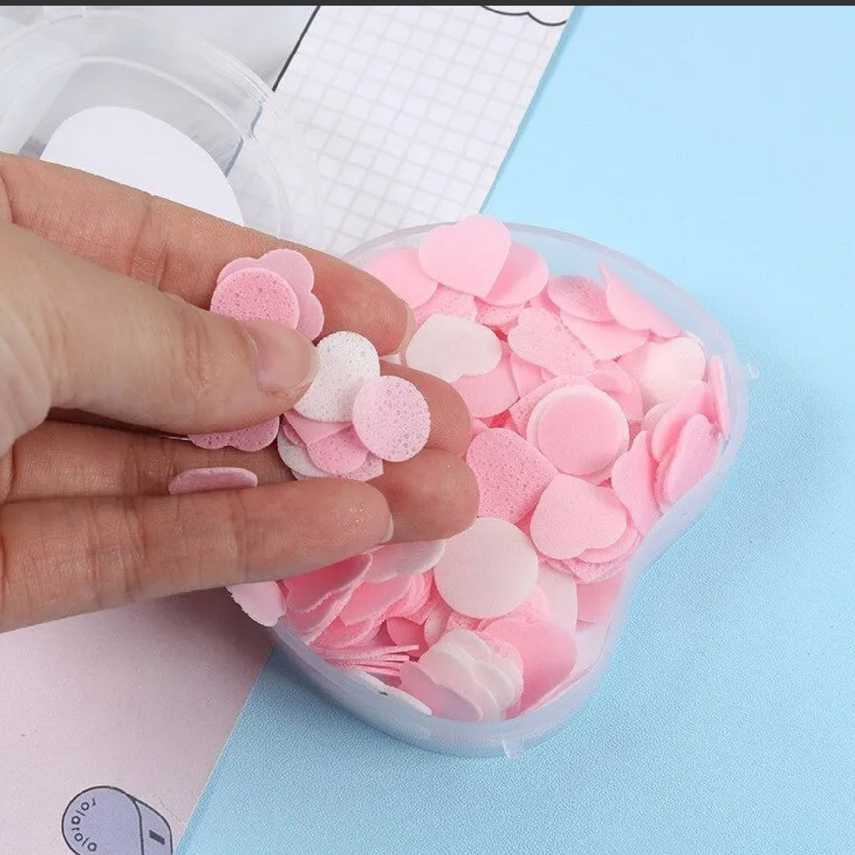 A hand holds several pink and white heart and circle-shaped paper soap pieces above an open apple-shaped container.
