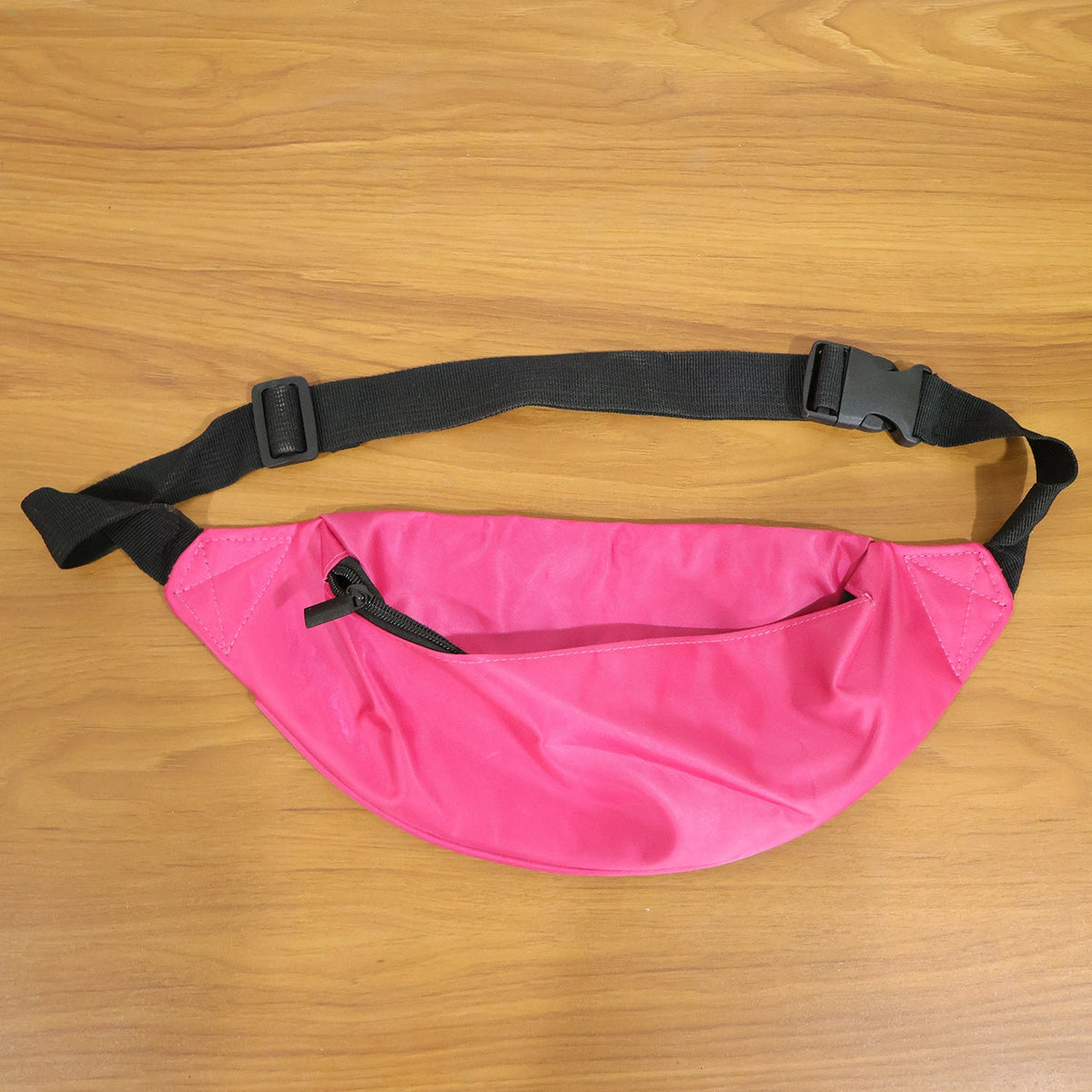 A bright pink fanny pack with an open front zipper and black strap rests on a wooden table.