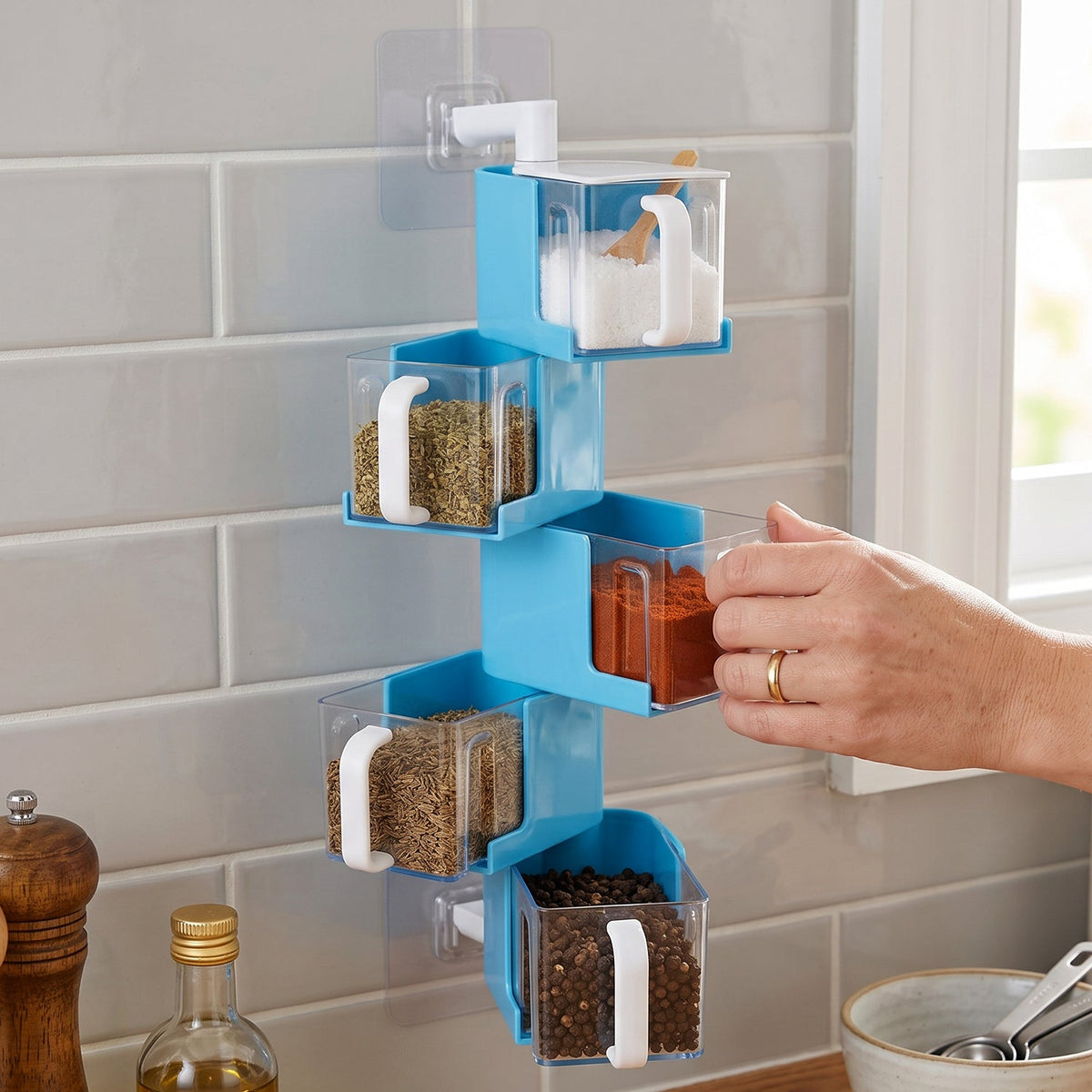 A hand pulls out a clear container of red spice from a blue 5-layer rotating spice rack on a tiled wall.