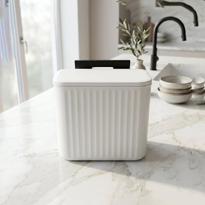 A white ribbed compost bin with a closed lid and black mounting bracket sits on a white marble kitchen counter.