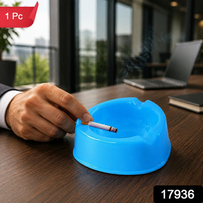 A hand holds a lit cigarette over a blue plastic ashtray on a wooden desk in an office setting.