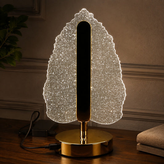 Illuminated crystal leaf lamp with a gold base and stem, plugged in on a dark wooden surface.