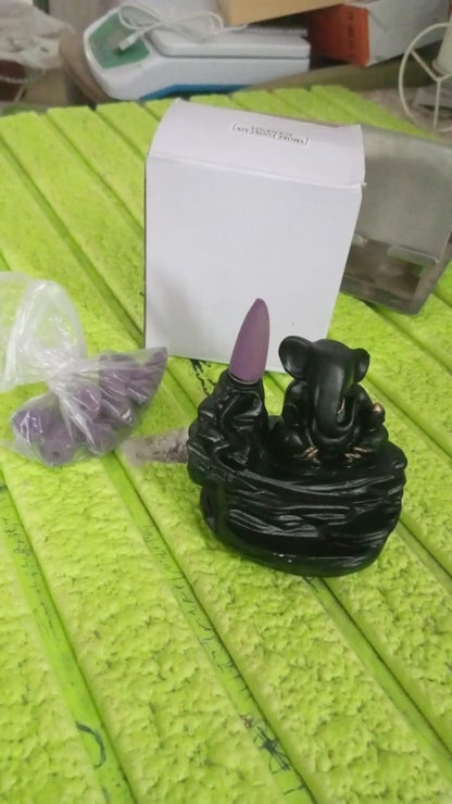 Black Ganesha smoke fountain with a purple incense cone, a white box, and a bag of purple cones on a textured green surface.