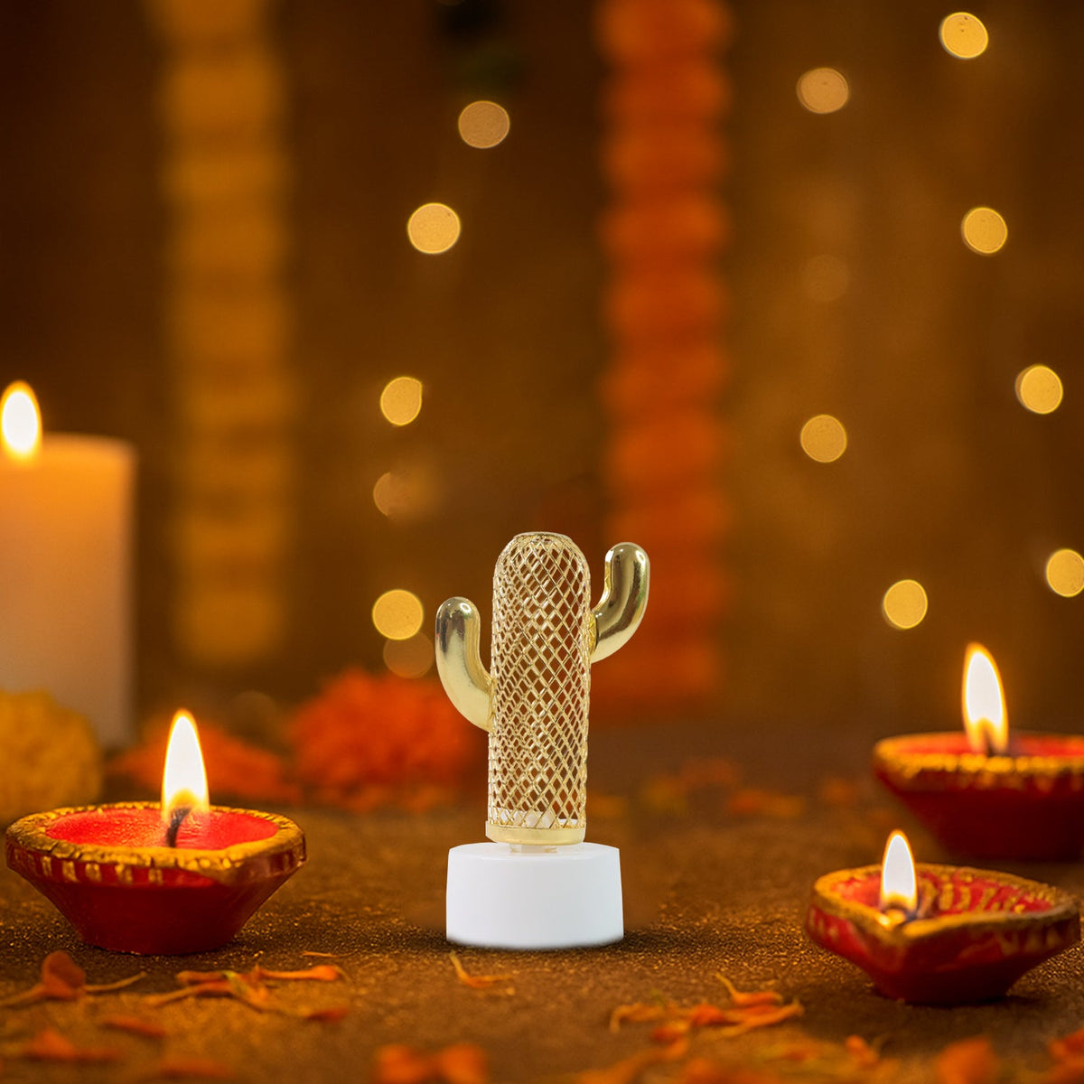 A single glowing cactus-shaped LED light surrounded by three lit traditional clay diyas and flower petals on a dark surface.