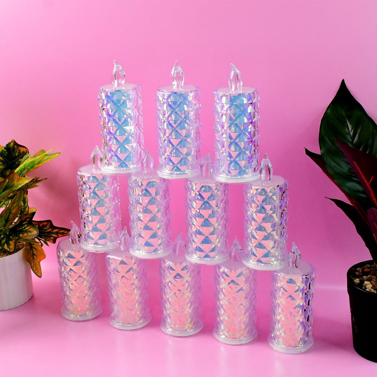 Twelve iridescent LED candles are stacked in a pyramid on a pink surface, with green and red plants on either side.