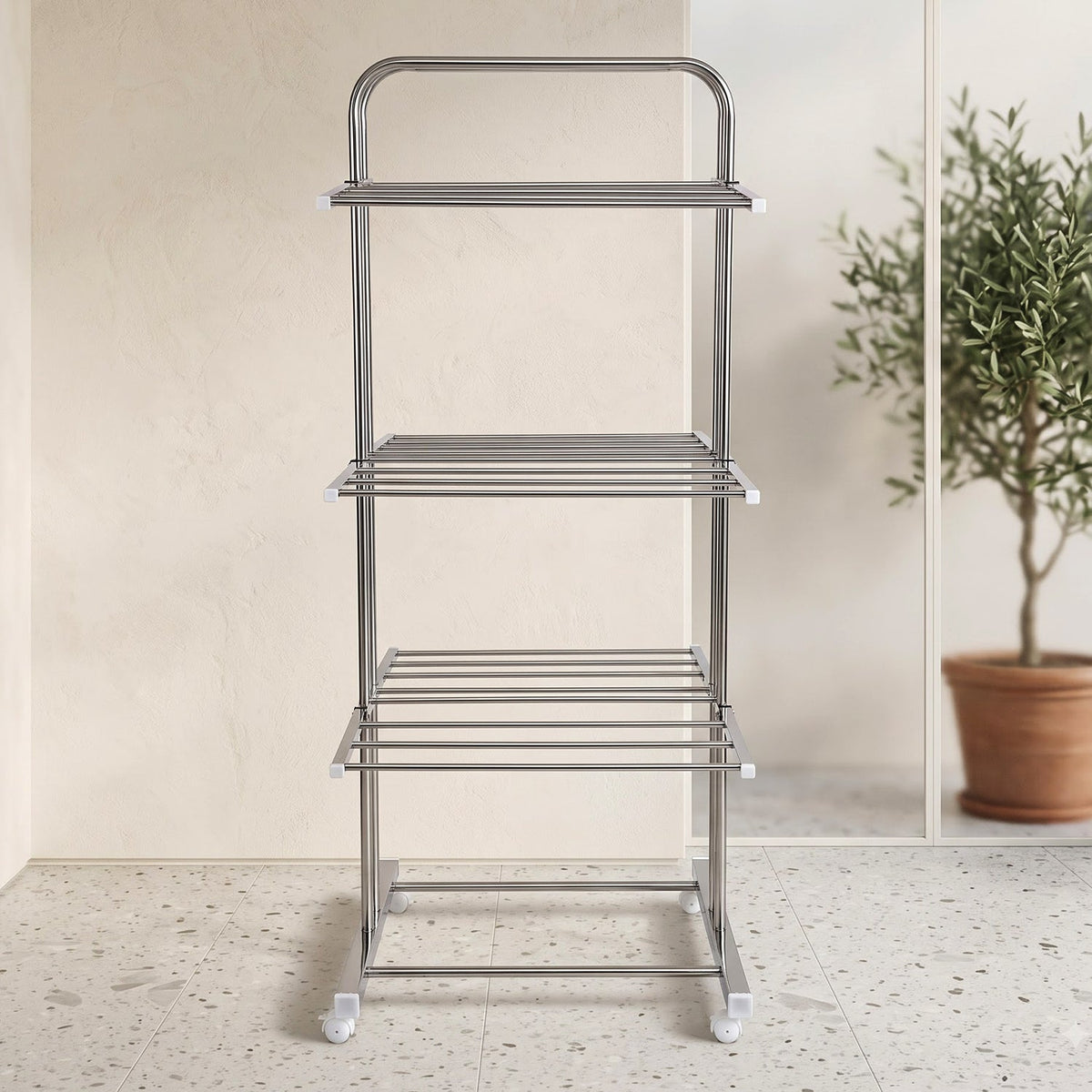 An empty stainless steel 3-layer clothes drying rack with wheels, in a bright room with a potted olive tree visible through