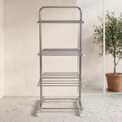 An empty stainless steel 3-layer clothes drying rack with wheels, in a bright room with a potted olive tree visible through