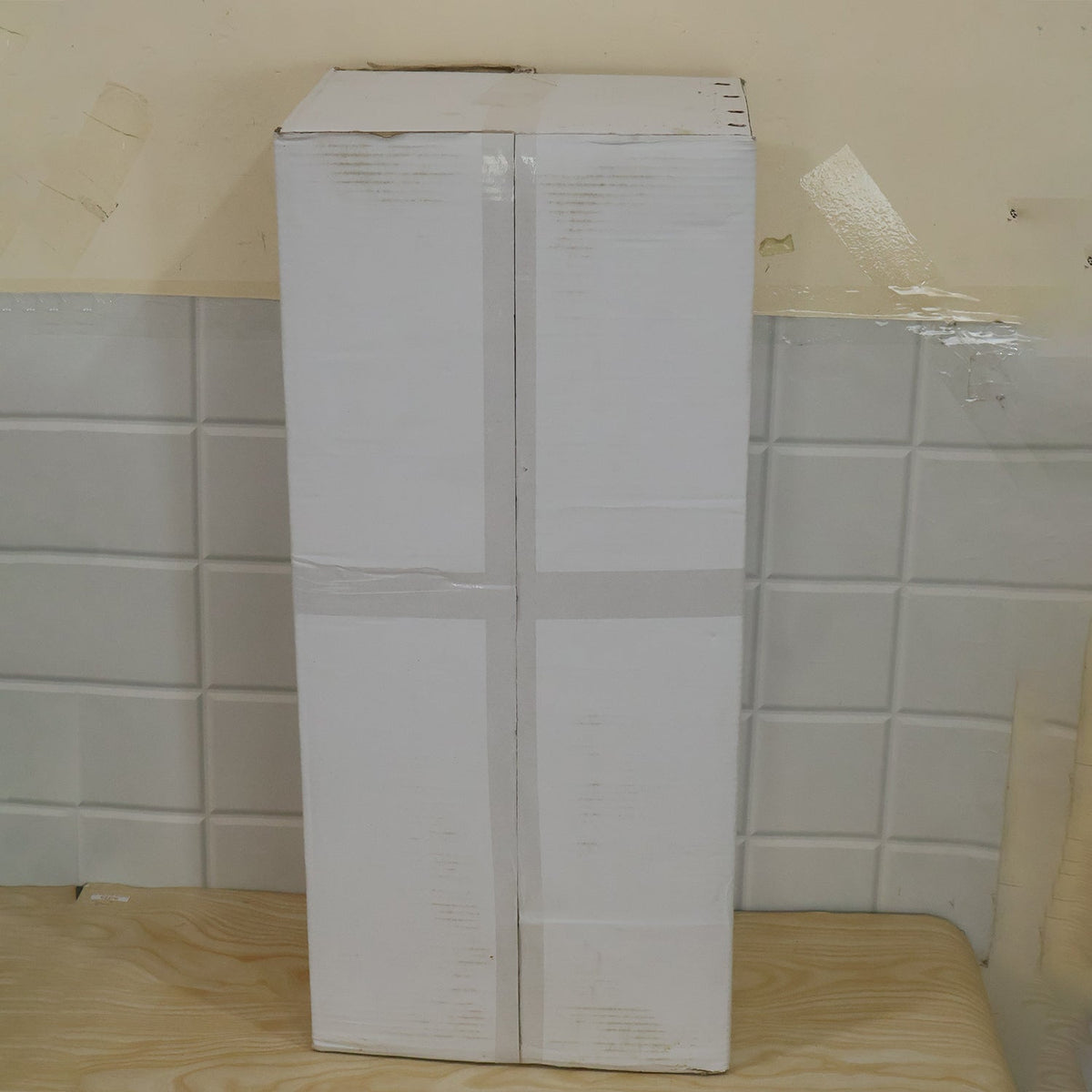 A tall, rectangular cardboard box sealed with tape, resting on a light wooden surface against a white tiled wall.