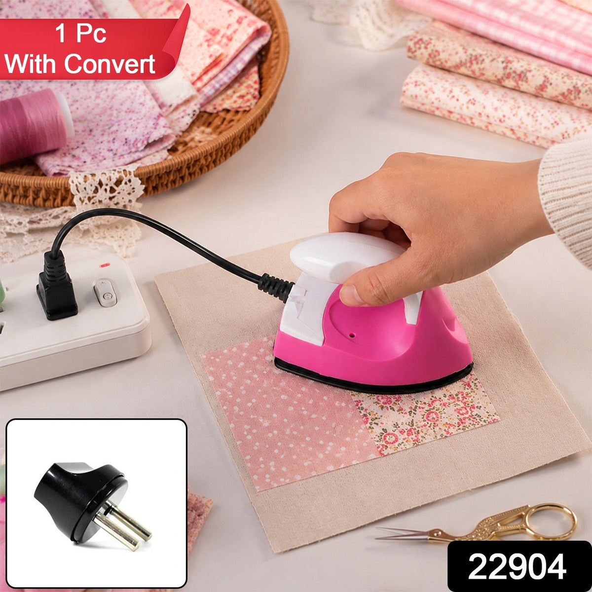 A hand holds a pink and white mini iron over a piece of floral fabric on a beige cloth, with a power strip, craft supplies,
