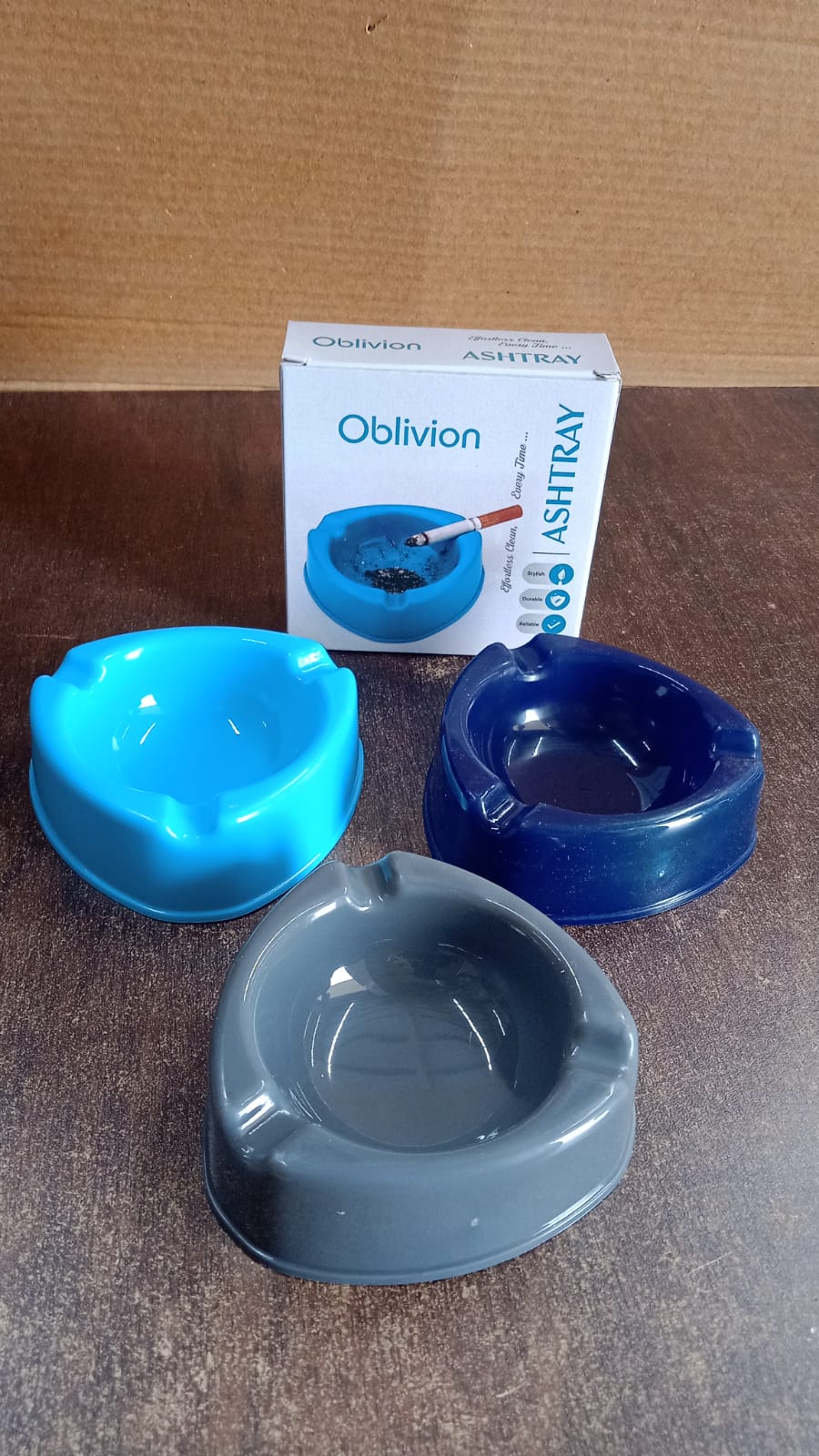 Three plastic ashtrays, one light blue, one dark blue, and one grey, are displayed with a product box in the background.