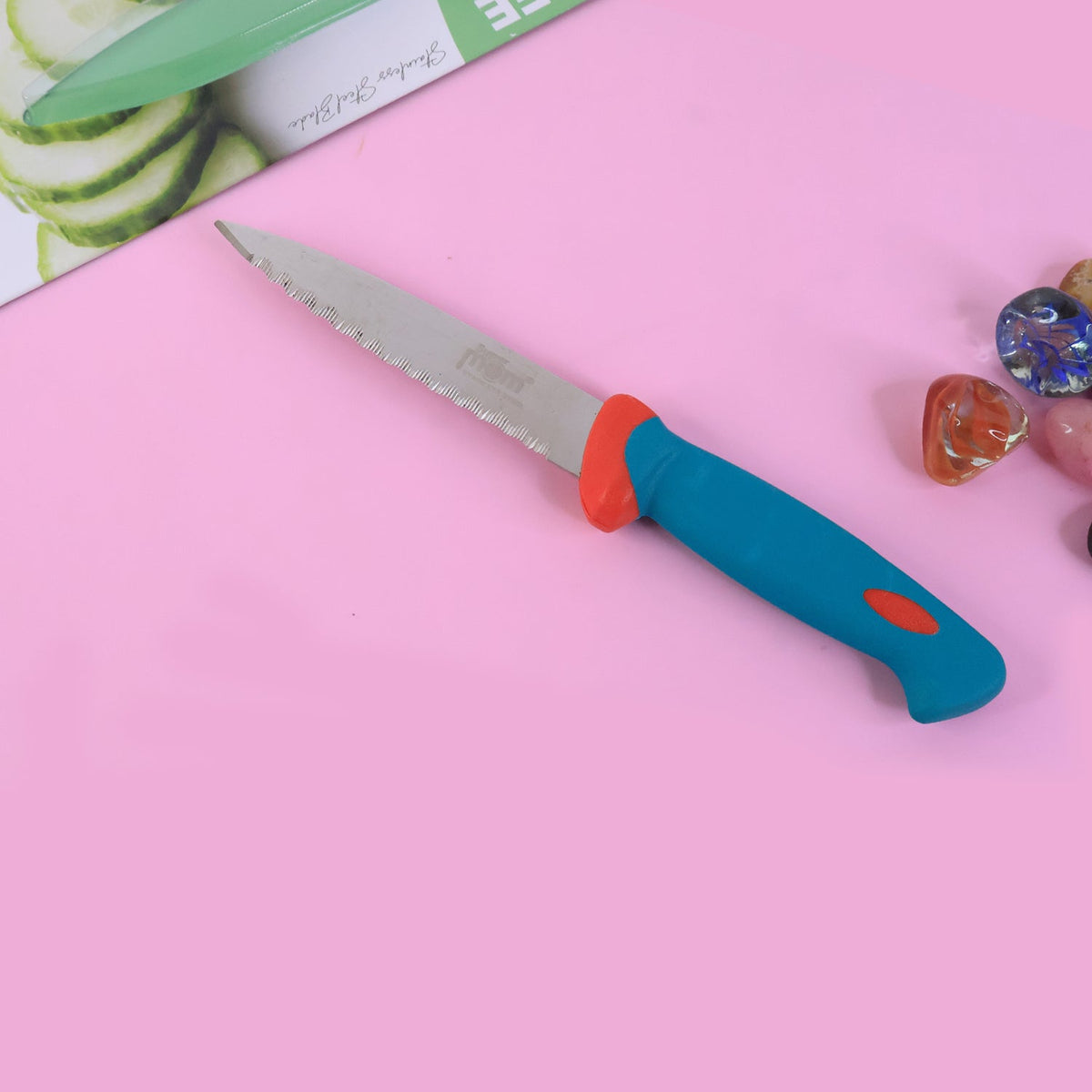 A blue and red handled serrated paring knife is positioned on a vibrant pink surface, with a blurred image of sliced