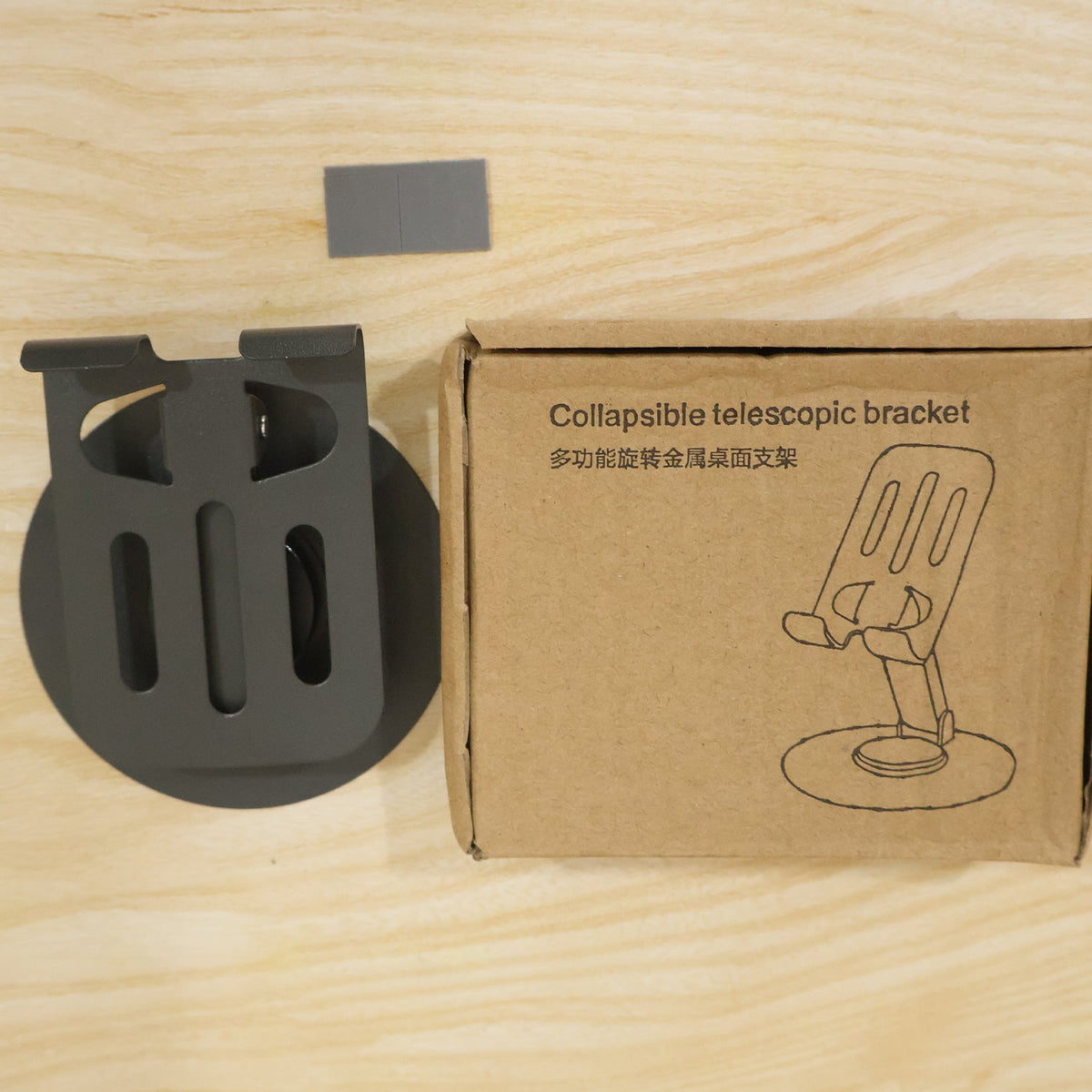 A black metal mobile stand next to its brown cardboard box labeled "Collapsible telescopic bracket" on a wooden surface.