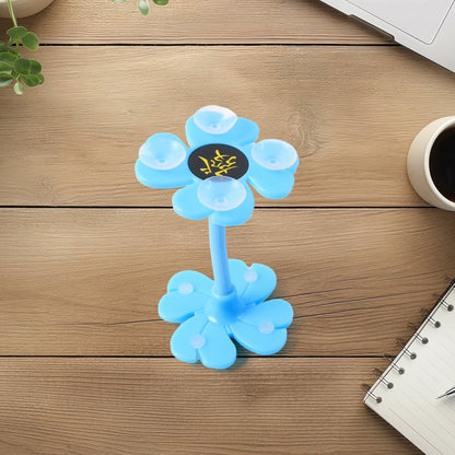 A blue flower-shaped phone holder with suction cups sits on a wooden desk with a notebook and coffee cup.