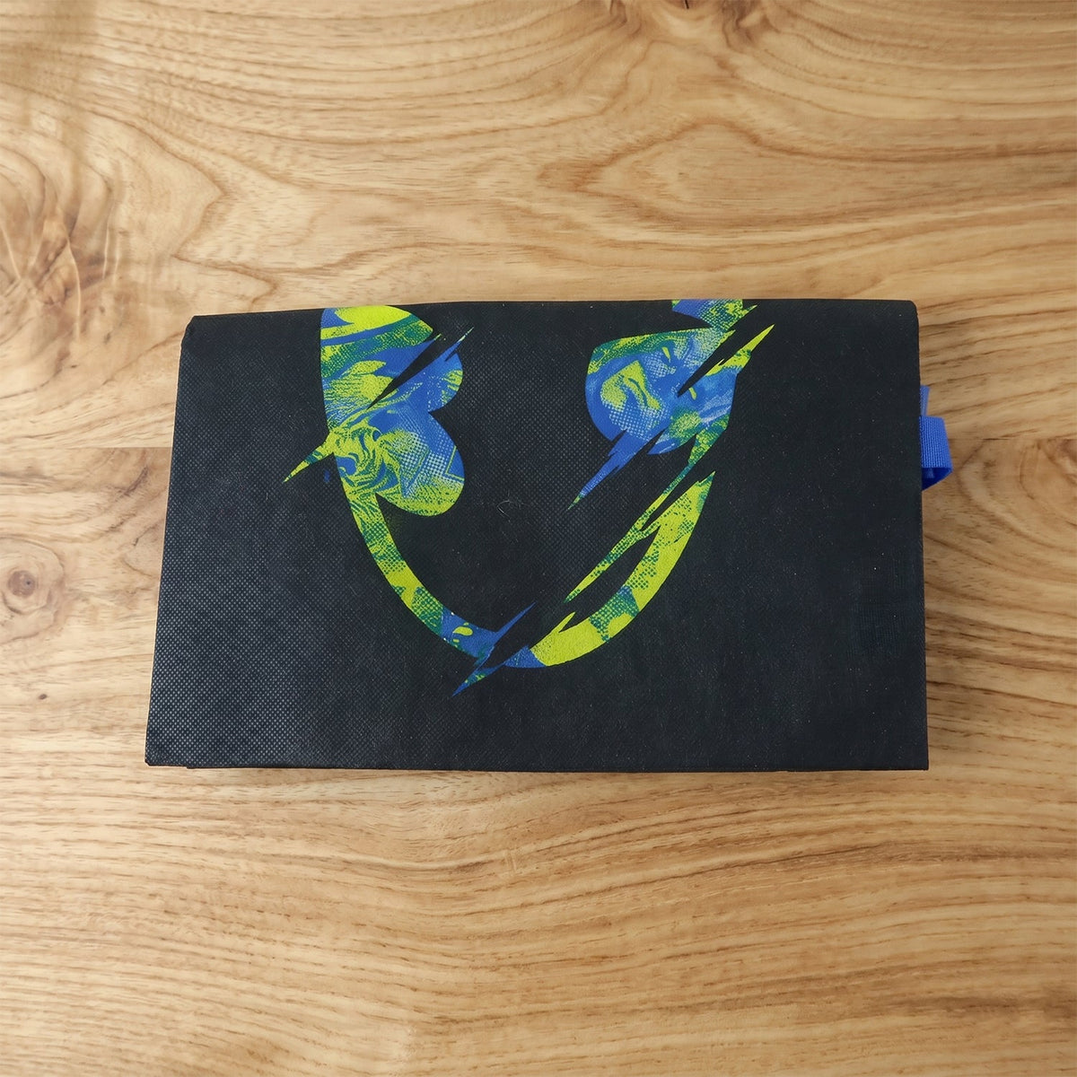 Folded black reusable tote bag with a distressed neon green and blue Batman logo on a wooden surface.