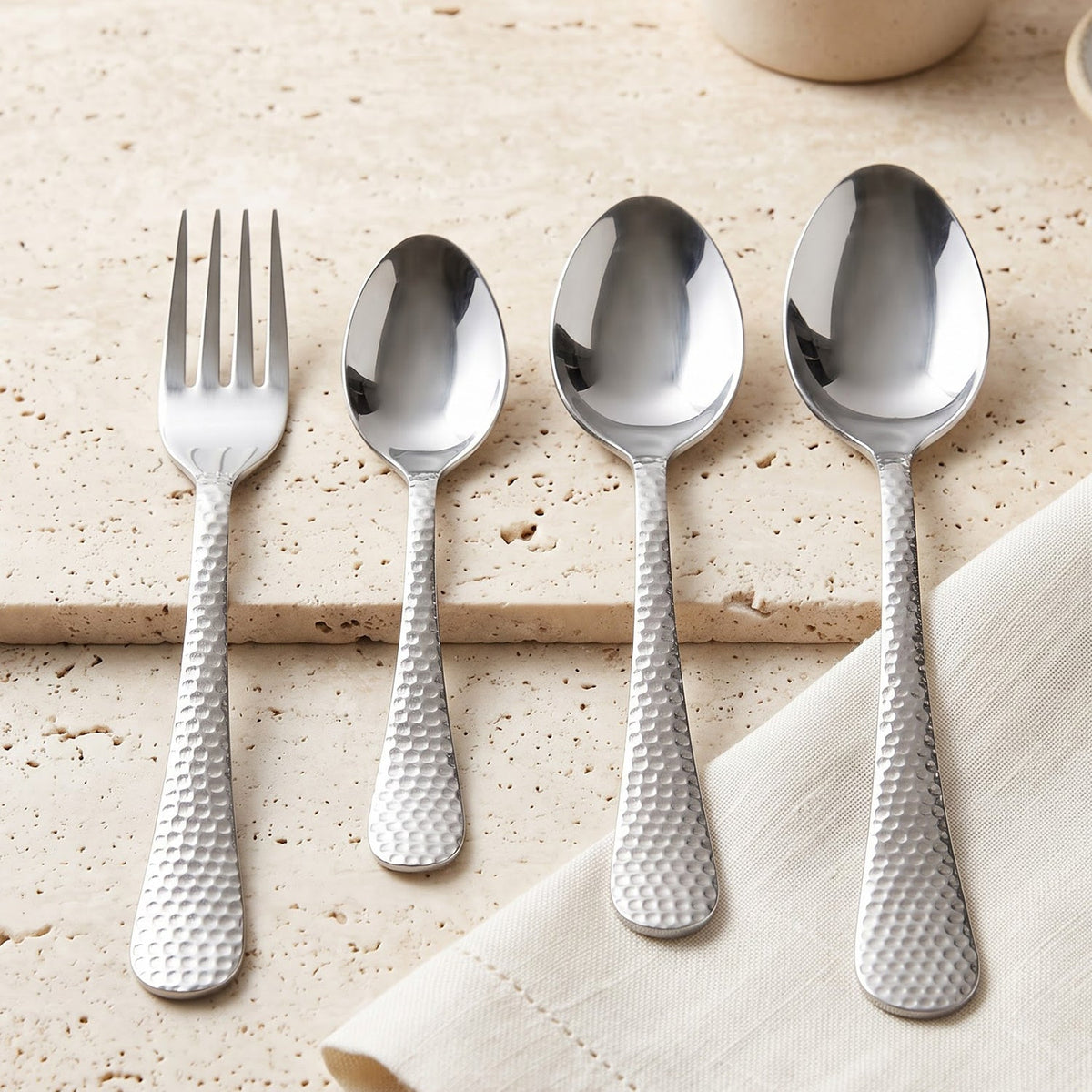 Four pieces of hammered stainless steel cutlery, including a fork, small spoon, and two larger spoons, are arranged on a
