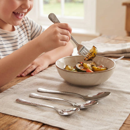 A smiling child uses a hammered stainless steel fork to eat roasted vegetables from a bowl. Three other pieces of cutlery