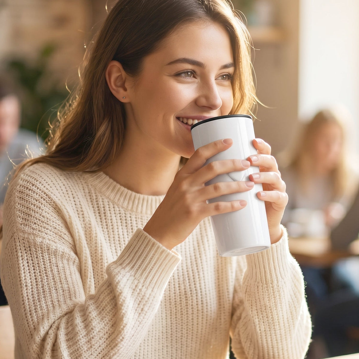 A smiling woman in a cream sweater holds a white thermal tumbler with a black rim in a cafe setting.