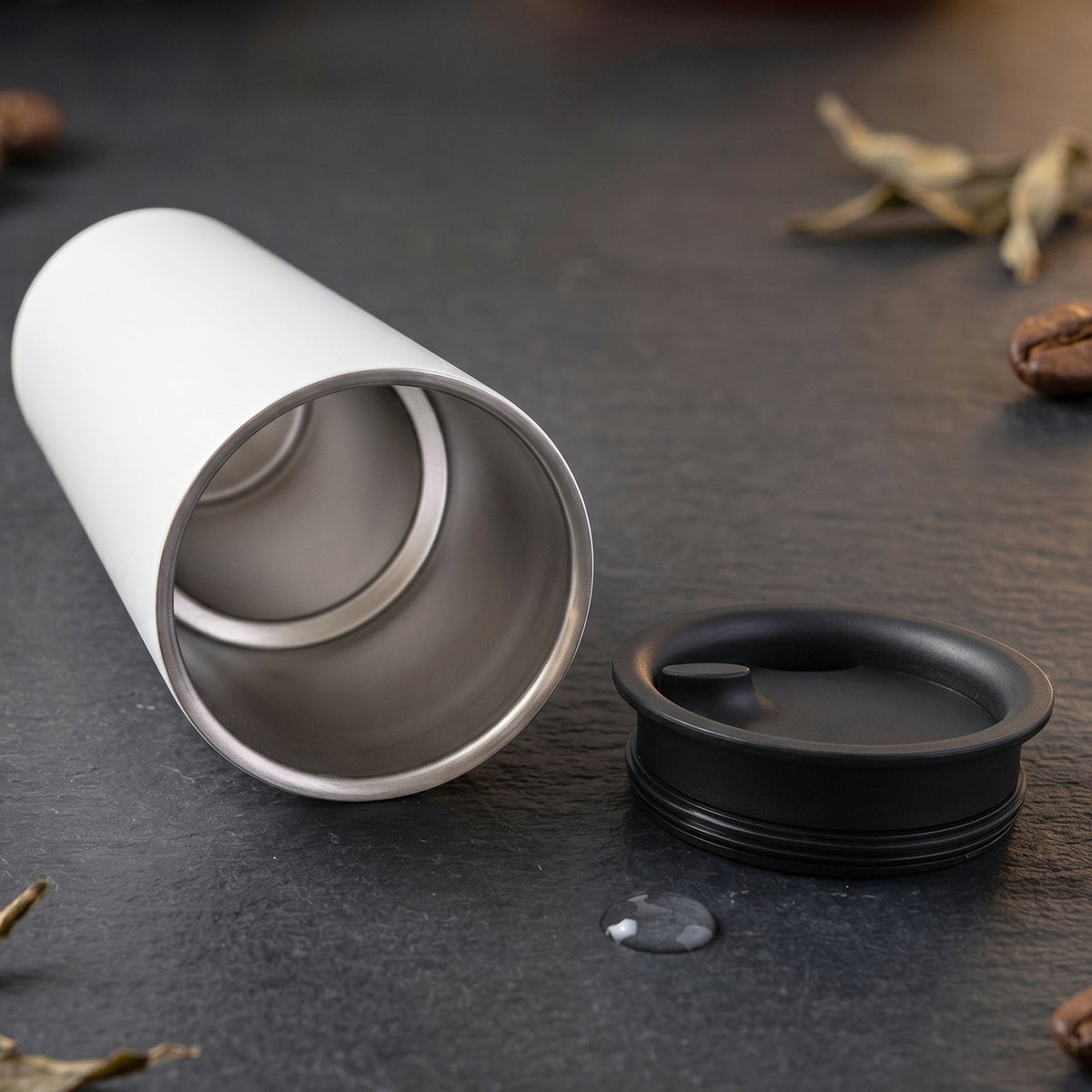 An open white thermal tumbler with a stainless steel interior and its black lid resting on a dark surface.