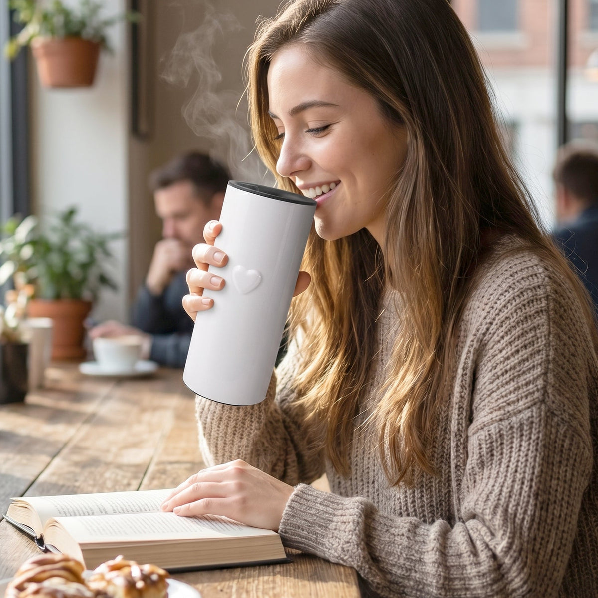 A woman sips from a white thermal tumbler with a heart detail, steam rising, while reading a book in a cafe.