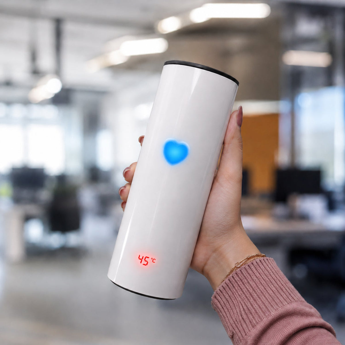 A hand holds a white thermal tumbler with a glowing blue heart and a red temperature display in an office.