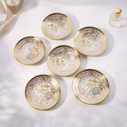 Six pink and gold floral porcelain saucers with gold rims and floral designs are arranged on a white surface.